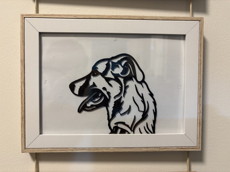 El retrato del perro impreso en negro, en un marco de madera clara con paspartú blanco, colgado de dos cuerdas en una pared beige.